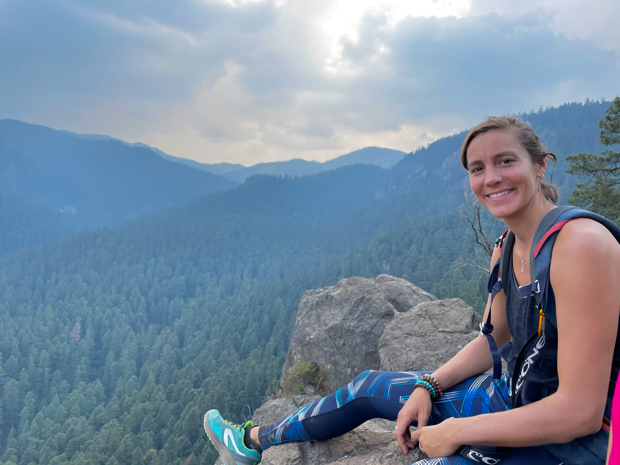 Femme souriante assise sur un sommet de montagne, entourée de montagnes, symbolisant le lâcher-prise, la connexion à soi, l'harmonie avec la nature et le sentiment d'accomplissement. Sophrologue Nice, inspirant le bien-être.