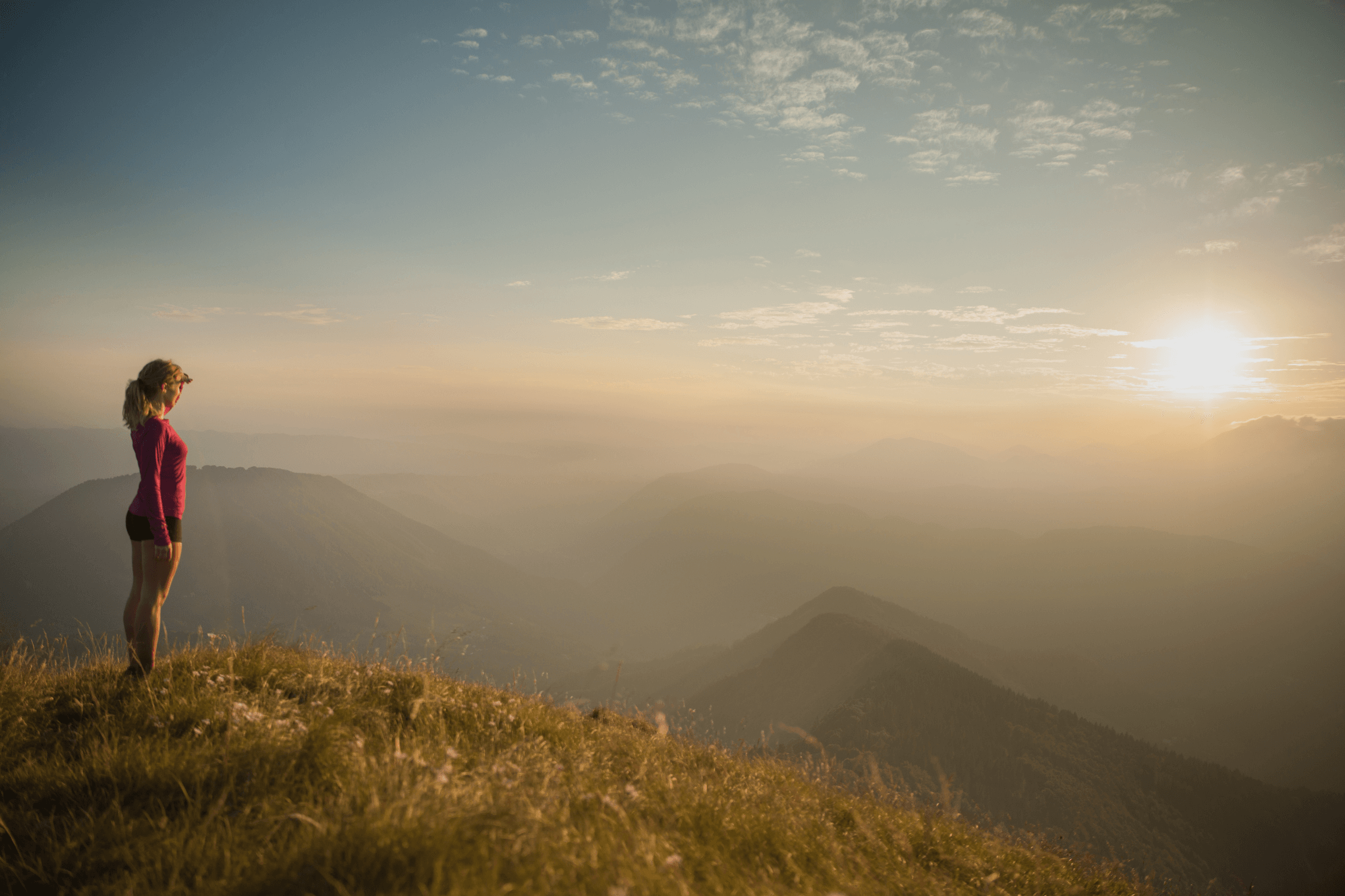 Femme debout sur un sommet de montagne, observant le coucher du soleil, symbolisant l'équilibre, la gestion du stress, la relaxation et le lâcher-prise dans un moment de zenitude.