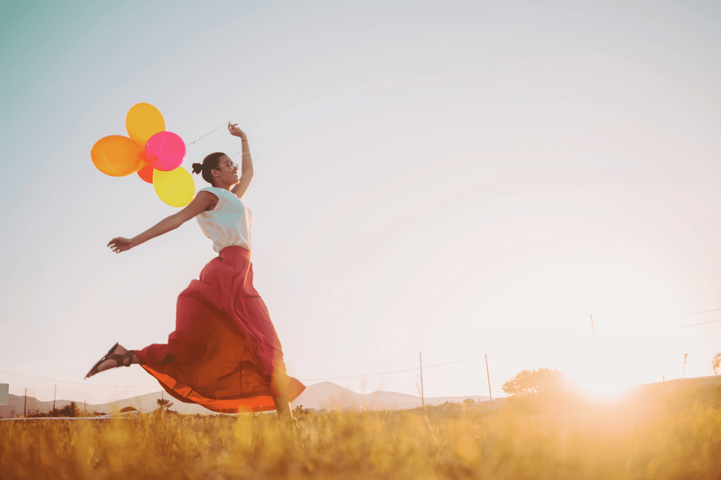 Femme dansant avec des ballons au coucher du soleil, symbolisant le lâcher prise, la connexion à soi et le moment présent. Lié aux bénéfices de la sophrologie et de la respiration pour un bien-être profond.